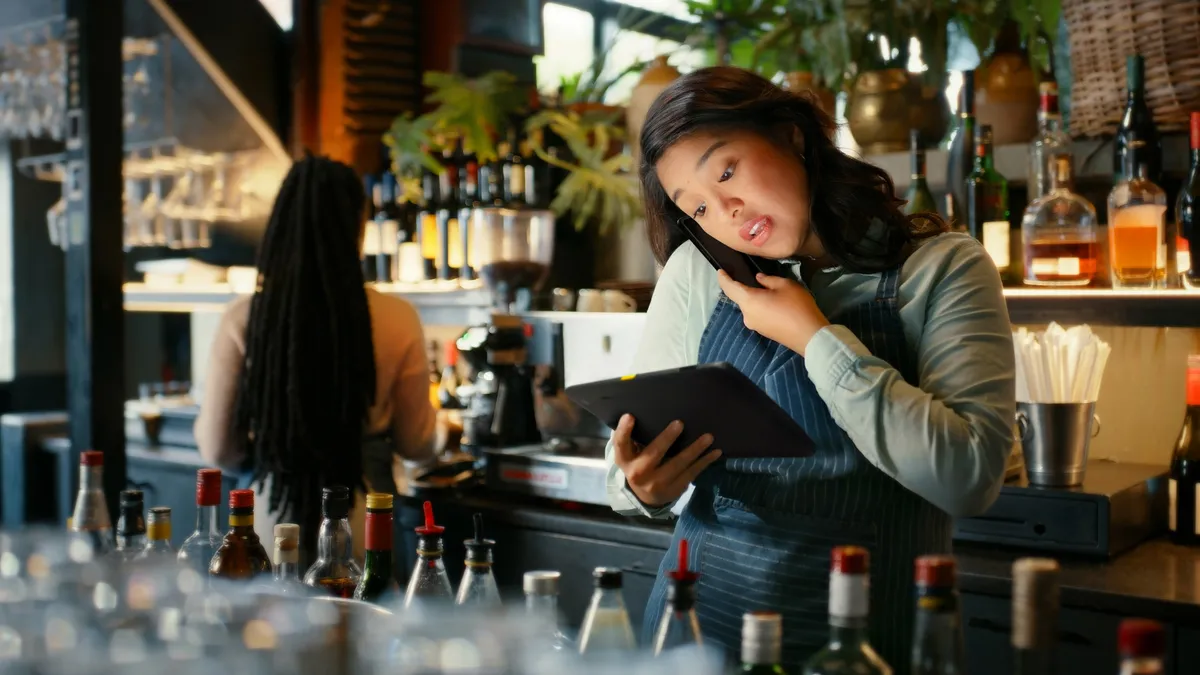 Atendente de restaurante no balcão segura a tablet com uma mão e o celular no ombro com a outra, durante o movimento do salão