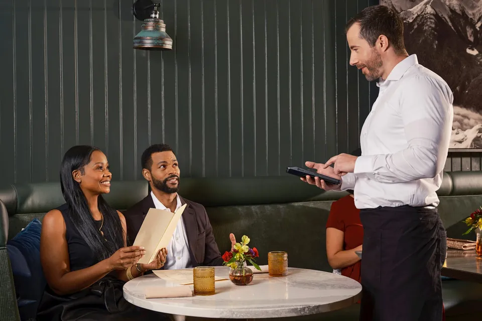 Garçom anota o pedido na mesa usando um POS de mão, com clientes escolhendo no menu